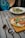 A table setting with a wooden surface holds a flatbread topped with melted cheese, arugula, and slices of cured meat. Next to it are stylish sunglasses. A vibrant blue cocktail with fruit floats nearby, and in the background, a bowl of pasta with clams and greens is partially visible.