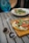 A table setting with a wooden surface holds a flatbread topped with melted cheese, arugula, and slices of cured meat. Next to it are stylish sunglasses. A vibrant blue cocktail with fruit floats nearby, and in the background, a bowl of pasta with clams and greens is partially visible.