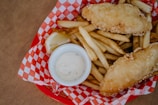 A red basket lined with checkered paper holds crispy golden-brown fish fillets and French fries. A small white bowl containing creamy dipping sauce and a slice of lemon is nestled among the fries.
