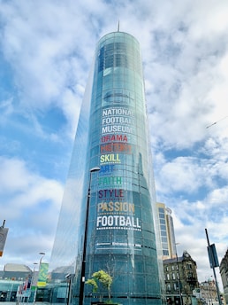 A modern glass building with the words 'National Football Museum' and various other terms like 'Drama', 'History', 'Skill', 'Art', 'Faith', 'Style', and 'Passion' written in colorful letters on its facade. The structure is situated against a backdrop of a partly cloudy sky with several lampposts and other buildings visible at street level.