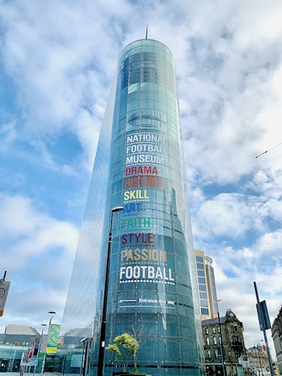 A modern glass building with the words 'National Football Museum' and various other terms like 'Drama', 'History', 'Skill', 'Art', 'Faith', 'Style', and 'Passion' written in colorful letters on its facade. The structure is situated against a backdrop of a partly cloudy sky with several lampposts and other buildings visible at street level.
