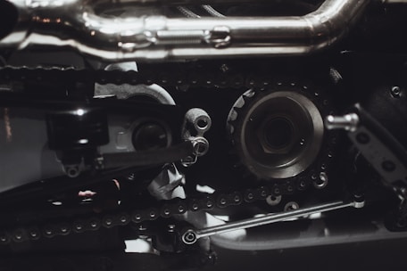 Intricate mechanical components featuring gears, chains, and metal parts, likely part of an engine or machinery. The image highlights metallic textures and detailed craftsmanship.