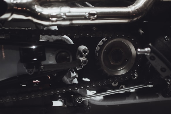 Intricate mechanical components featuring gears, chains, and metal parts, likely part of an engine or machinery. The image highlights metallic textures and detailed craftsmanship.