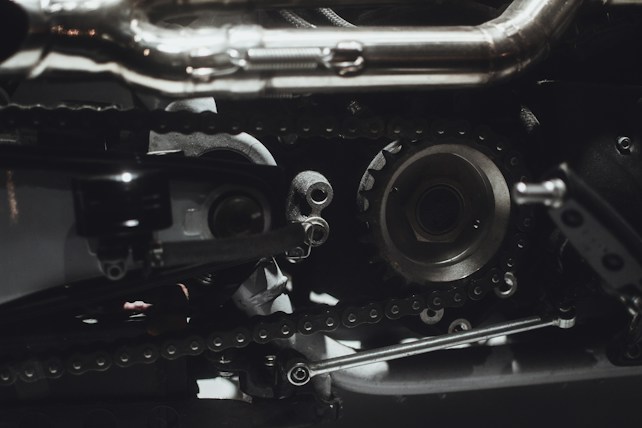 Intricate mechanical components featuring gears, chains, and metal parts, likely part of an engine or machinery. The image highlights metallic textures and detailed craftsmanship.