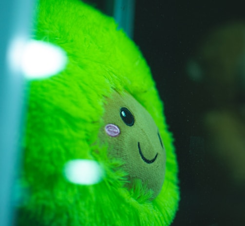A close-up of a plush toy with vibrant green fur and a smiling face, pressed against a glass surface. The toy has rosy cheeks and is displayed in a glass enclosure or a vending machine.