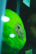 A close-up of a plush toy with vibrant green fur and a smiling face, pressed against a glass surface. The toy has rosy cheeks and is displayed in a glass enclosure or a vending machine.