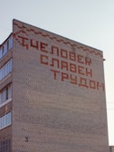 A multi-story brick building with an inscription in large Cyrillic letters on its facade. The text is arranged decoratively across a blank section of the wall. Several windows are visible on the side of the building.