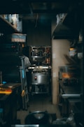 A narrow, dimly lit kitchen space with various cooking equipment and utensils neatly arranged. The area includes stainless steel appliances, shelves with containers, and a metal ladder leaning against the wall. A person in a white chef jacket is seen in the background focused on their task.