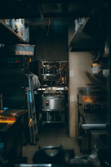 A narrow, dimly lit kitchen space with various cooking equipment and utensils neatly arranged. The area includes stainless steel appliances, shelves with containers, and a metal ladder leaning against the wall. A person in a white chef jacket is seen in the background focused on their task.