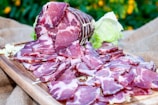 Slices of cured meat arranged on a wooden board with a backdrop of greenery and scattered lettuce leaves. The cured meat is deep red with marbled fat, and the board rests on a burlap cloth.