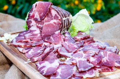 Slices of cured meat arranged on a wooden board with a backdrop of greenery and scattered lettuce leaves. The cured meat is deep red with marbled fat, and the board rests on a burlap cloth.