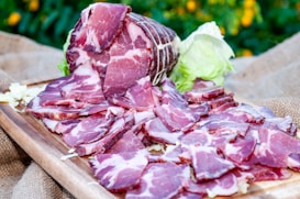 Slices of cured meat arranged on a wooden board with a backdrop of greenery and scattered lettuce leaves. The cured meat is deep red with marbled fat, and the board rests on a burlap cloth.
