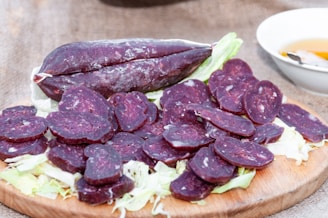 Sliced dark purple sausage placed on a wooden board, garnished with fresh lettuce leaves. A whole sausage is also shown, placed lengthwise. A white bowl with a sauce and a spoon is in the background on a textured surface.
