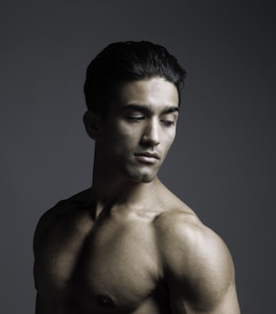 A shirtless male figure with a muscular build poses against a dark, subdued background. His head is tilted downward, and his gaze is directed away from the camera, creating a contemplative mood. The lighting accentuates the contours of his body, highlighting muscular definition.