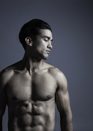 A shirtless man with a muscular physique stands against a dark gray background. His head is slightly turned to the side, eyes closed, exhibiting a calm and contemplative expression. The lighting emphasizes the contours of his body, highlighting his well-defined muscles.
