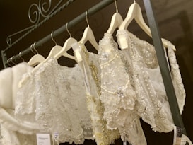 A collection of ornate white and ivory garments, possibly wedding dresses or formal attire, is displayed on hangers. The garments are heavily embellished with beads, sequins, lace, and intricate embroidery. The rack is metallic and the hangers are uniform in style.