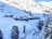 A snowy alpine landscape with a cluster of wooden cabins nestled at the base of a mountain. Snow-covered slopes and pine trees surround the area, creating a serene and picturesque scene. The bright sunlight casts shadows on the hillside, adding depth to the tranquil setting.