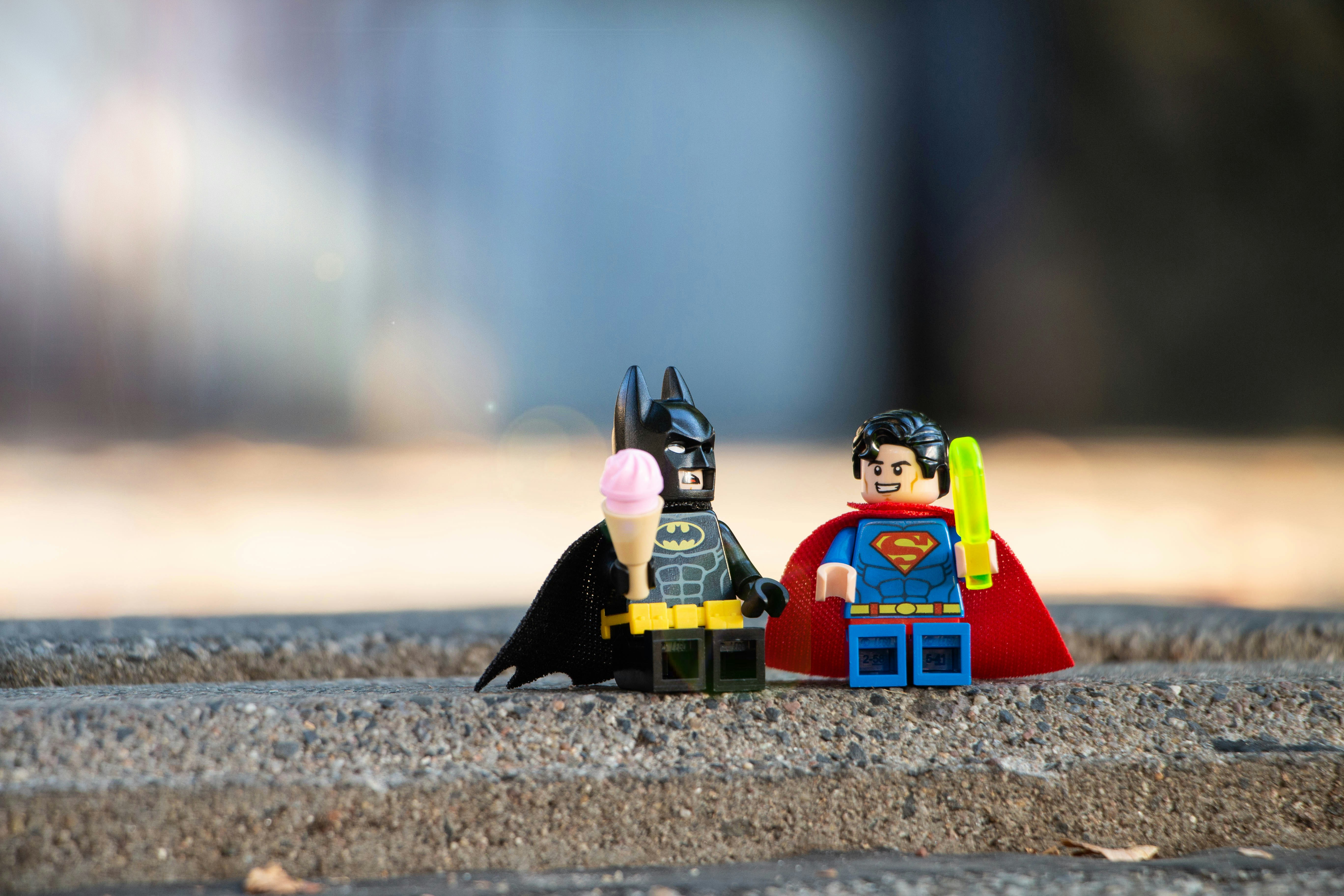 A Lego minifigure of Batman holding an icecream next to a Lego minifigure of Superman holding a sword