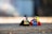 Two LEGO figures of superhero characters are standing on a textured surface. One figure resembles a character in a black costume with a pink cone accessory, while the other is dressed in blue and red with a green accessory. The background is blurred, creating a bokeh effect.