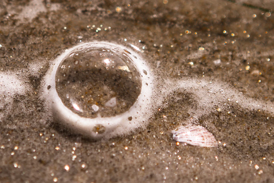 Round, clear bubble floats on sandy beach with surrounding smaller white bubbles nearby
