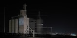 A large industrial building stands in the darkness, illuminated by sparse artificial lighting. The structure is labeled with 'Bolton's Crown Quality' and 'Feed & Seed Co. Quality Supreme'. The scene features multiple silos and a train with several freight cars on the tracks. Power lines crisscross above the building, adding an industrial vibe to the setting.