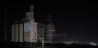 A large industrial building stands in the darkness, illuminated by sparse artificial lighting. The structure is labeled with 'Bolton's Crown Quality' and 'Feed & Seed Co. Quality Supreme'. The scene features multiple silos and a train with several freight cars on the tracks. Power lines crisscross above the building, adding an industrial vibe to the setting.