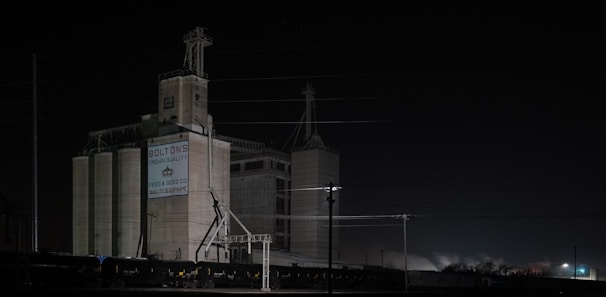 A large industrial building stands in the darkness, illuminated by sparse artificial lighting. The structure is labeled with 'Bolton's Crown Quality' and 'Feed & Seed Co. Quality Supreme'. The scene features multiple silos and a train with several freight cars on the tracks. Power lines crisscross above the building, adding an industrial vibe to the setting.