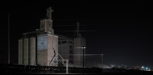 A large industrial building stands in the darkness, illuminated by sparse artificial lighting. The structure is labeled with 'Bolton's Crown Quality' and 'Feed & Seed Co. Quality Supreme'. The scene features multiple silos and a train with several freight cars on the tracks. Power lines crisscross above the building, adding an industrial vibe to the setting.
