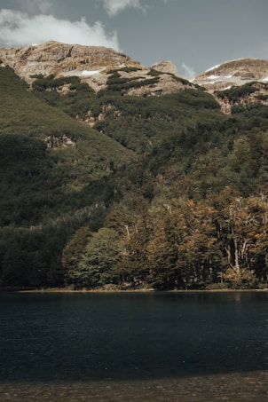 A serene mountain landscape features lush green forests at the base, with towering rocky peaks in the background. A tranquil body of water occupies the foreground, reflecting the natural scenery above. The sky is mostly clear with a few clouds, enhancing the peaceful atmosphere.