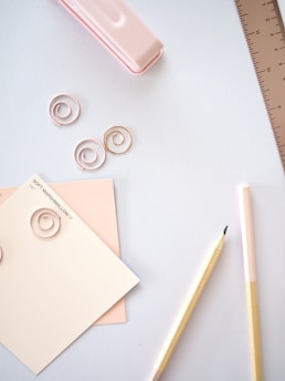 A pastel-themed stationery flat lay including a pink stapler, spiral paper clips, paint swatches labeled as 'Soft Marshmallow', a wooden pencil, and a light pink ruler on a white surface.