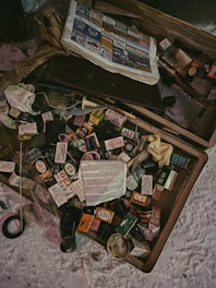 An assortment of scattered, vintage film boxes and booklets is spread out on a flat surface. A newspaper with colorful advertisements is partially visible in the upper section. Various small objects, possibly related to photography, are interspersed among the film boxes.