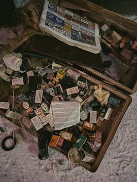 An assortment of scattered, vintage film boxes and booklets is spread out on a flat surface. A newspaper with colorful advertisements is partially visible in the upper section. Various small objects, possibly related to photography, are interspersed among the film boxes.