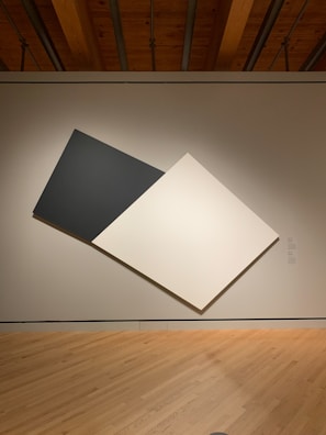 A minimalist art piece displayed on a gallery wall. The artwork consists of two large, overlapping geometric panels, one black and one white, positioned diagonally against a neutral background. The room features wooden floors and a ceiling with visible beams.