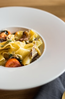 A plate of pasta garnished with sliced cherry tomatoes and herbs, presented on a wide white plate with a minimalistic design. The pasta appears to be freshly cooked, with a rich combination of yellow and golden hues.