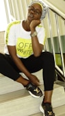 A person wearing a white T-shirt with bright yellow stripes and the words OFF-WHITE printed on the front, black pants, and stylish black sneakers with a 'LV' logo. They are sitting on indoor stairs, leaning their head on one hand, and wearing a wristwatch and glasses.