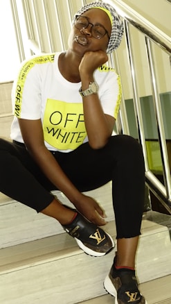 A person wearing a white T-shirt with bright yellow stripes and the words OFF-WHITE printed on the front, black pants, and stylish black sneakers with a 'LV' logo. They are sitting on indoor stairs, leaning their head on one hand, and wearing a wristwatch and glasses.