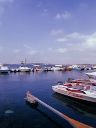 A harbor scene with multiple boats moored along a dock under a clear sky. The water appears calm, reflecting the blue of the sky. In the foreground, a speedboat prominently displaying the Yamaha brand is visible. A distant cityscape with a tall building and a flag can be seen across the water.