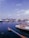 A harbor scene with multiple boats moored along a dock under a clear sky. The water appears calm, reflecting the blue of the sky. In the foreground, a speedboat prominently displaying the Yamaha brand is visible. A distant cityscape with a tall building and a flag can be seen across the water.