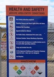 A prominently displayed safety instruction sign at a secure area entry point. It lists various rules including: port safety induction required, flashing beacon and headlights on, children not permitted, speed limit of 20km/h, no public entry, extreme caution with heavy machinery, no handheld devices, drug and alcohol-free site, specific clothing and footwear requirements, and no bicycles. Additionally, it warns of hazardous substances present, such as oxidizing and flammable agents, toxic and corrosive materials, with emergency contact information provided.