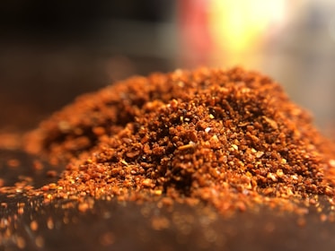 A close-up view of a pile of finely ground red chili powder spread across a surface. The texture is coarse, with visible flecks of bright red and orange colors. The image is lit in a way that highlights the granular texture and vibrant hues.