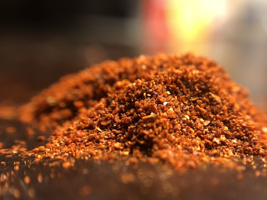 A close-up view of a pile of finely ground red chili powder spread across a surface. The texture is coarse, with visible flecks of bright red and orange colors. The image is lit in a way that highlights the granular texture and vibrant hues.