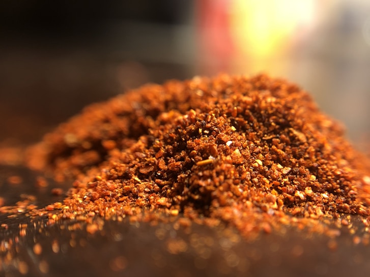 A close-up view of a pile of finely ground red chili powder spread across a surface. The texture is coarse, with visible flecks of bright red and orange colors. The image is lit in a way that highlights the granular texture and vibrant hues.