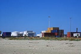 A series of stacked shipping containers in a variety of colors are positioned near a shoreline, with a sandy beach in the foreground. Behind the containers, large white storage tanks are visible under a clear blue sky. Sparse vegetation and a few tall light poles are also present.