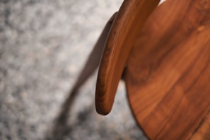 A close-up view of a wooden chair focusing on the curved backrest. The rich, natural wood grain is prominent, with smooth textures and a warm brown color. The background consists of a speckled, out-of-focus surface that contrasts with the chair's polished wood.