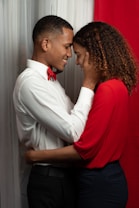 Two people are embracing closely, with their foreheads touching. One person is wearing a white shirt with a red bow tie, while the other has on a red top. They appear to be in front of red and white curtains, creating a warm and intimate setting.