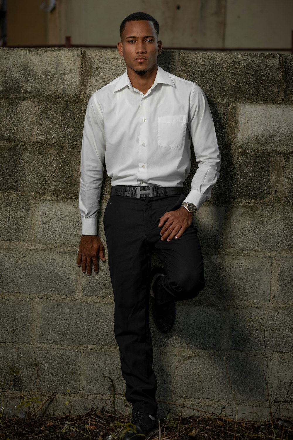 homem na camisa social branca e calças pretas apoiadas na parede de concreto cinza