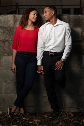 A man and a woman are standing close together, holding hands in front of a concrete wall. The woman is wearing a red blouse and dark pants while the man is dressed in a white shirt and dark trousers. They are looking into each other's eyes, with the woman smiling softly.