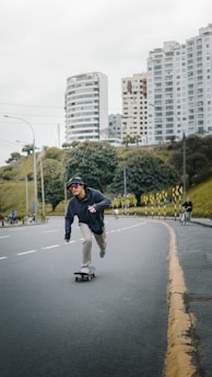 A person is skateboarding on a paved urban road while wearing casual attire and sunglasses. Tall residential buildings and greenery are visible in the background, creating an urban landscape. The road is marked with traffic lines and a yellow curb, and there are other people and cyclists in the distance.
