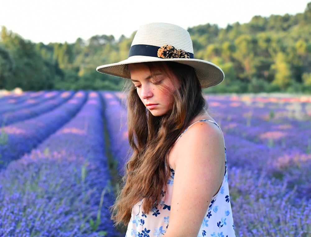 woman in blue and white floral sleeveless dress wearing brown hat standing on purple flower field