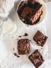 A plate filled with chocolate brownies is positioned next to a glass bottle of milk. Several brownies are scattered on a parchment paper, displaying a rich, crumbly texture.
